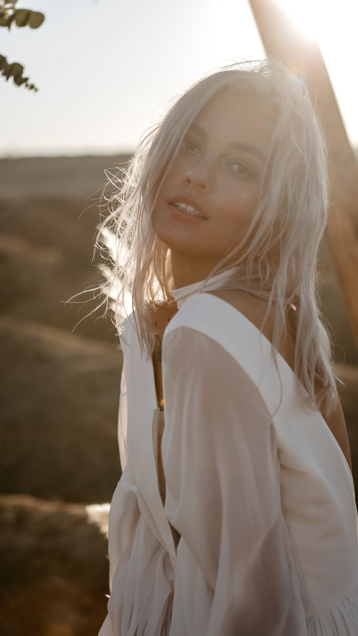 Sunlit portrait of a bride glancing back, sheer sleeve and wide V back of a chiffon sample wedding dress, wind in her hair, When the Moon is Out Sample