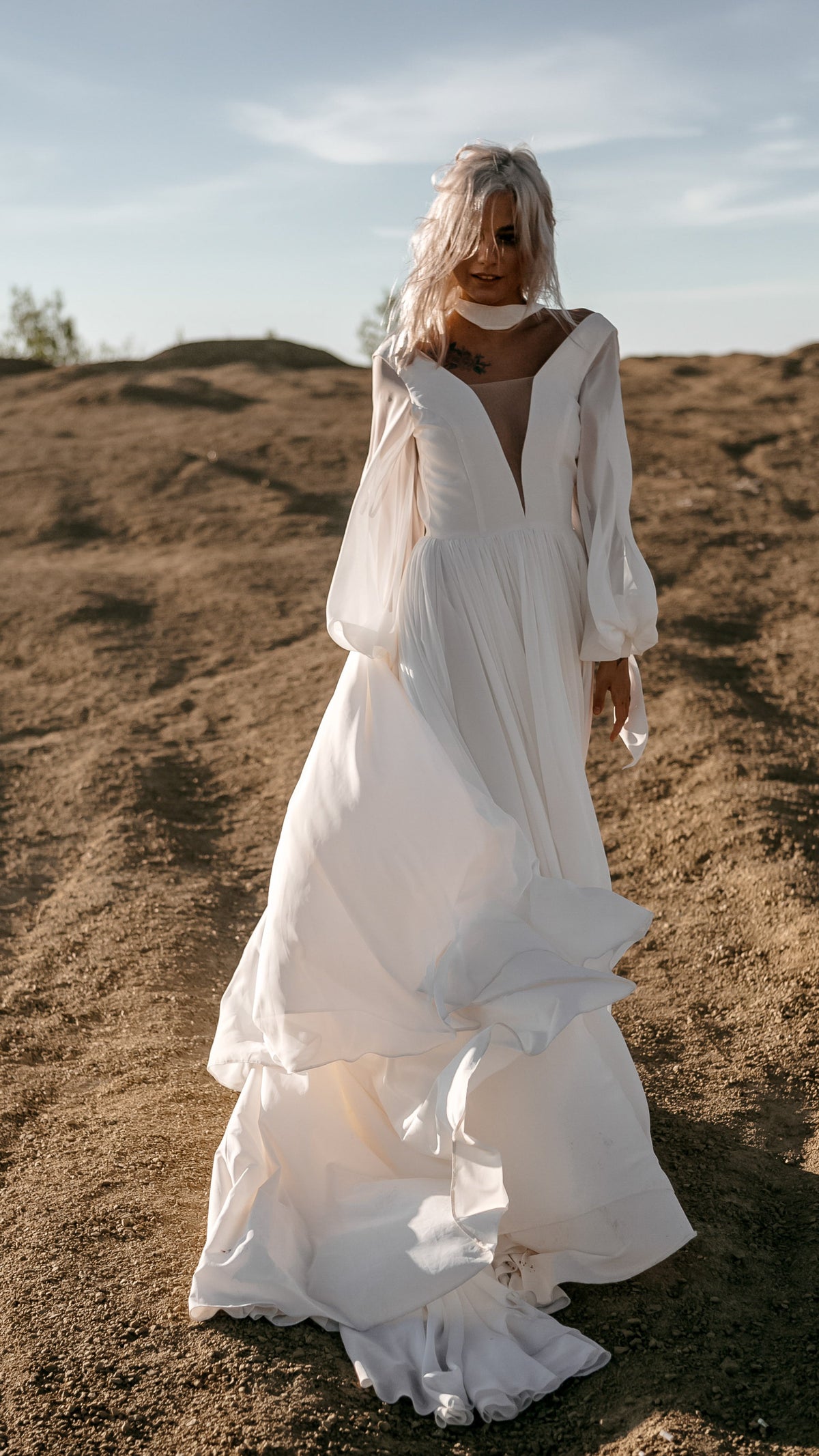 Bride walking toward the camera on a hill in a plunging chiffon sample wedding dress with long sleeves, the skirt lifting, When the Moon is Out Sample
