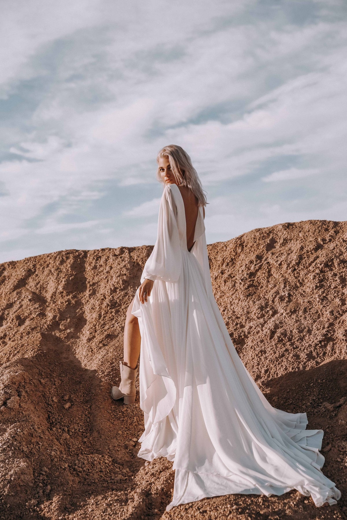 Bride looking back on a sandy ridge in a long-sleeve chiffon sample wedding dress, open back and train spilling down the slope, When the Moon is Out Sample