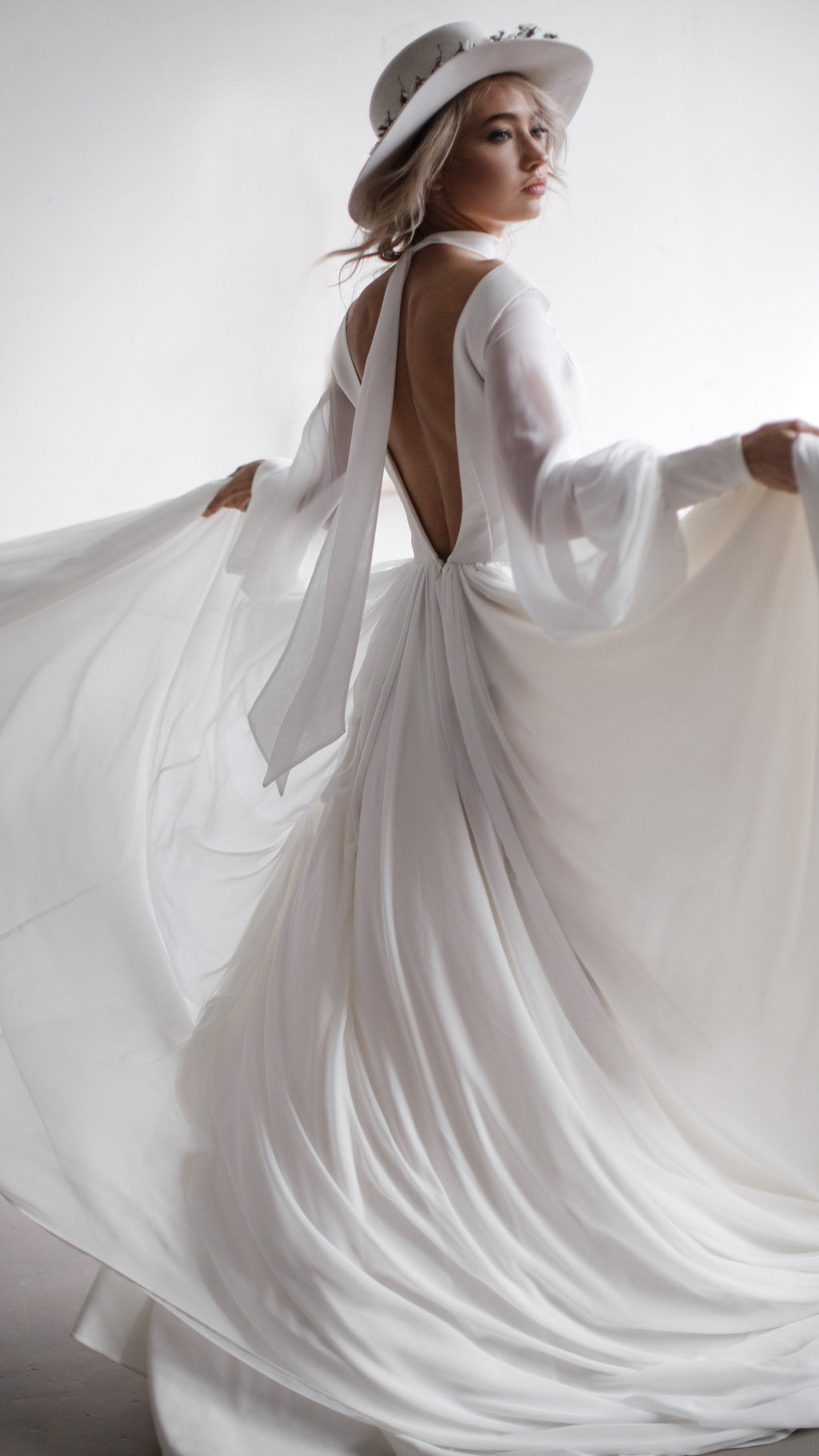 Bride turning in a hat, chiffon sample wedding dress with an open back and long sleeves, skirt held out in a white studio, When the Moon is Out Sample