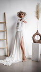 Bride leaning on a white wall in a long-sleeve chiffon wedding dress with a thigh slit and train, nude heels, When the Moon is Out