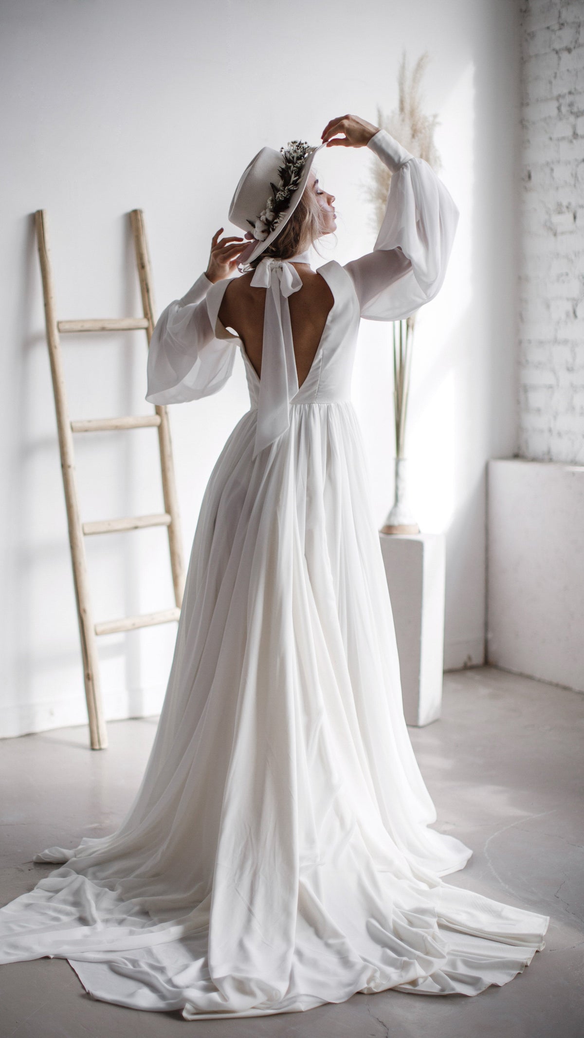 Back view of a bride adjusting her hat, keyhole open back and long chiffon sleeves of a wedding dress with a full train, When the Moon is Out