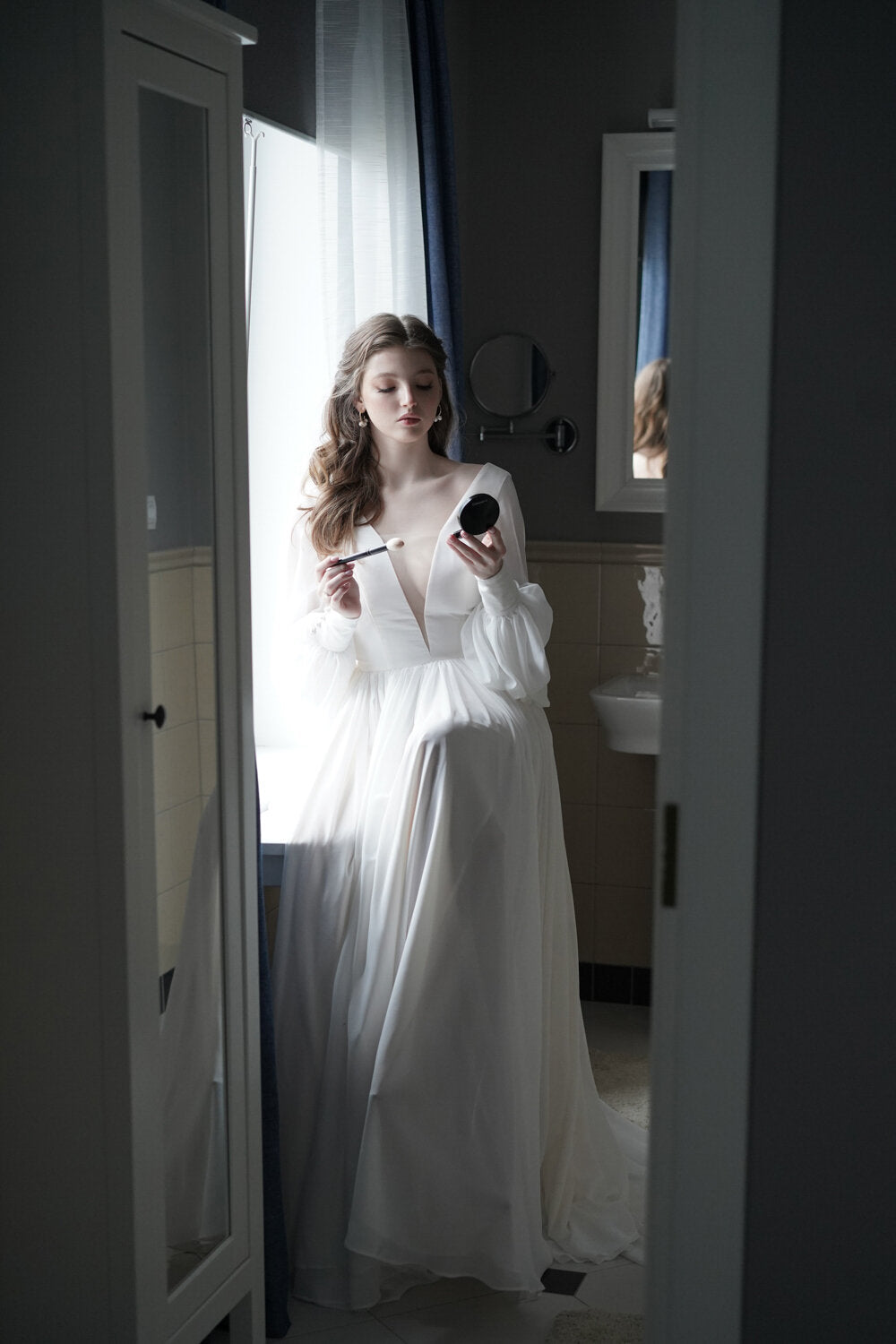 Bride getting ready by a bathroom mirror in a long-sleeve chiffon wedding dress with a plunging V neckline, When the Moon is Out