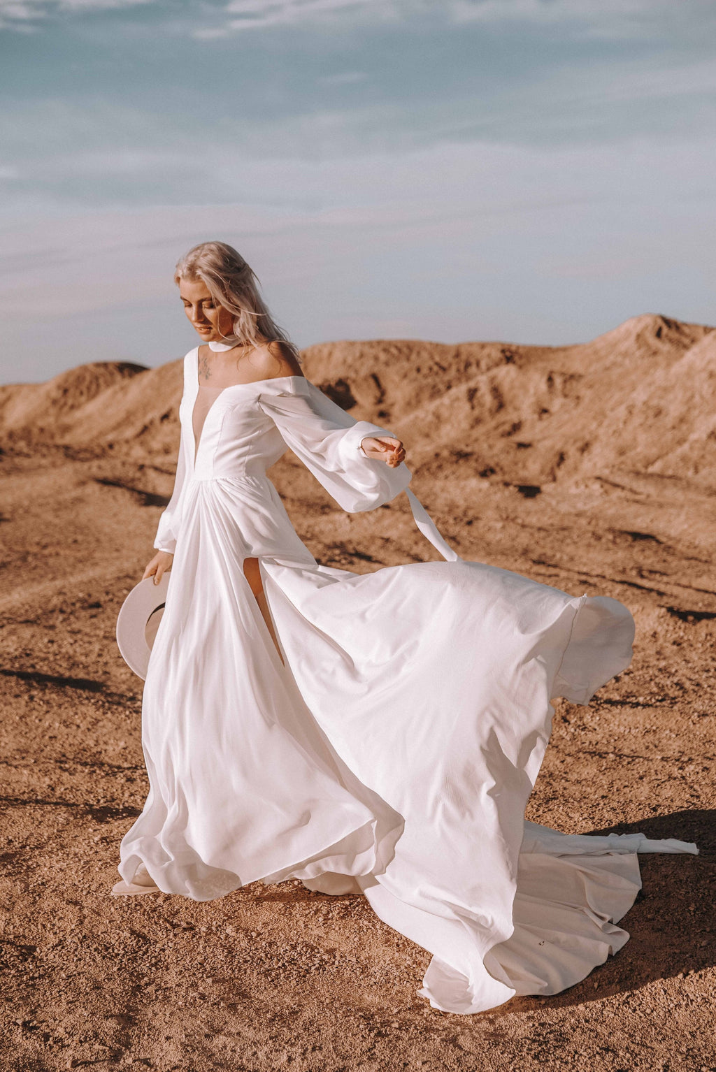Bride twirling on red dunes in a chiffon wedding dress with an off-shoulder V neckline and long sleeves, hat in hand, When the Moon is Out