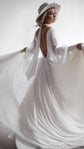 Bride turning in a hat, chiffon wedding dress with an open back and long sleeves, skirt held out in a white studio, When the Moon is Out