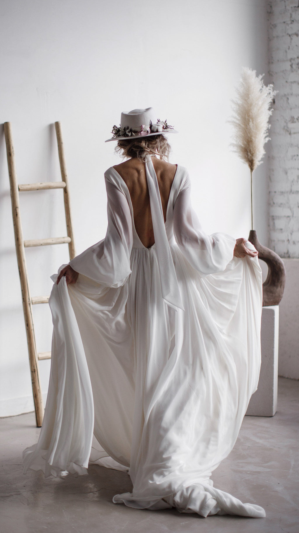 Back view of a bride lifting the chiffon skirt of a long-sleeve wedding dress, deep V back and train on the floor, When the Moon is Out