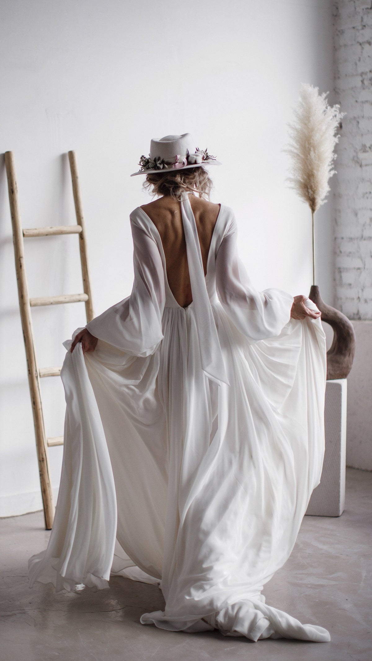 Back view of a bride lifting the chiffon skirt of a long-sleeve wedding dress, deep V back and train on the floor, When the Moon is Out