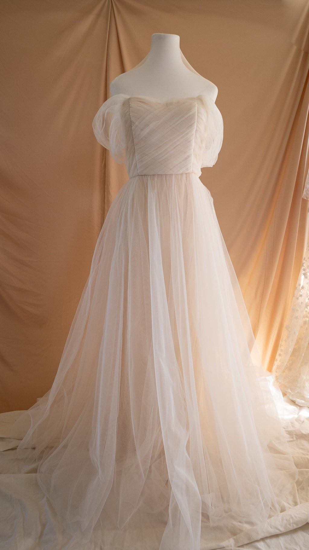 Champagne tulle sample wedding dress on a mannequin, pleated off-shoulder bodice with soft puff sleeves and a long train, Whisper Sample by Ephemeris
