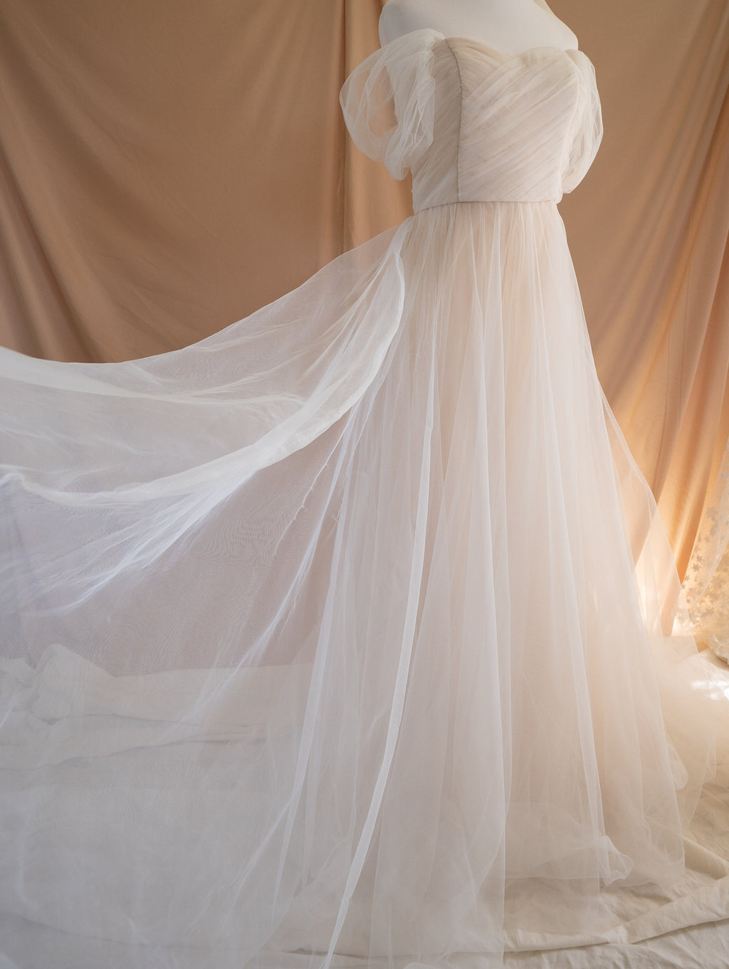Side view of a champagne tulle sample wedding dress on a mannequin with the layered train spread wide across the floor, Whisper Sample