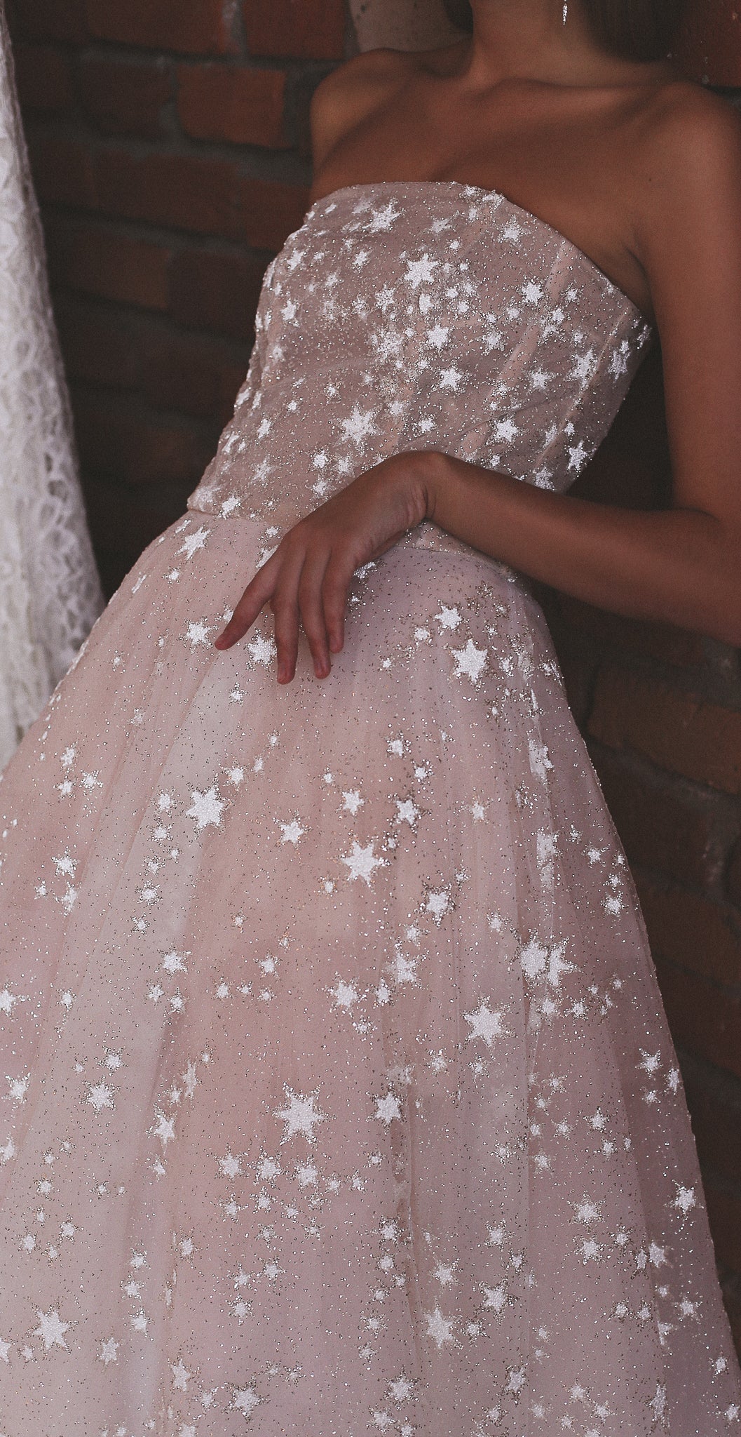 Crop of the strapless boned bodice of a star wedding dress, hand resting on the full tulle skirt, brick wall, Wild Stars