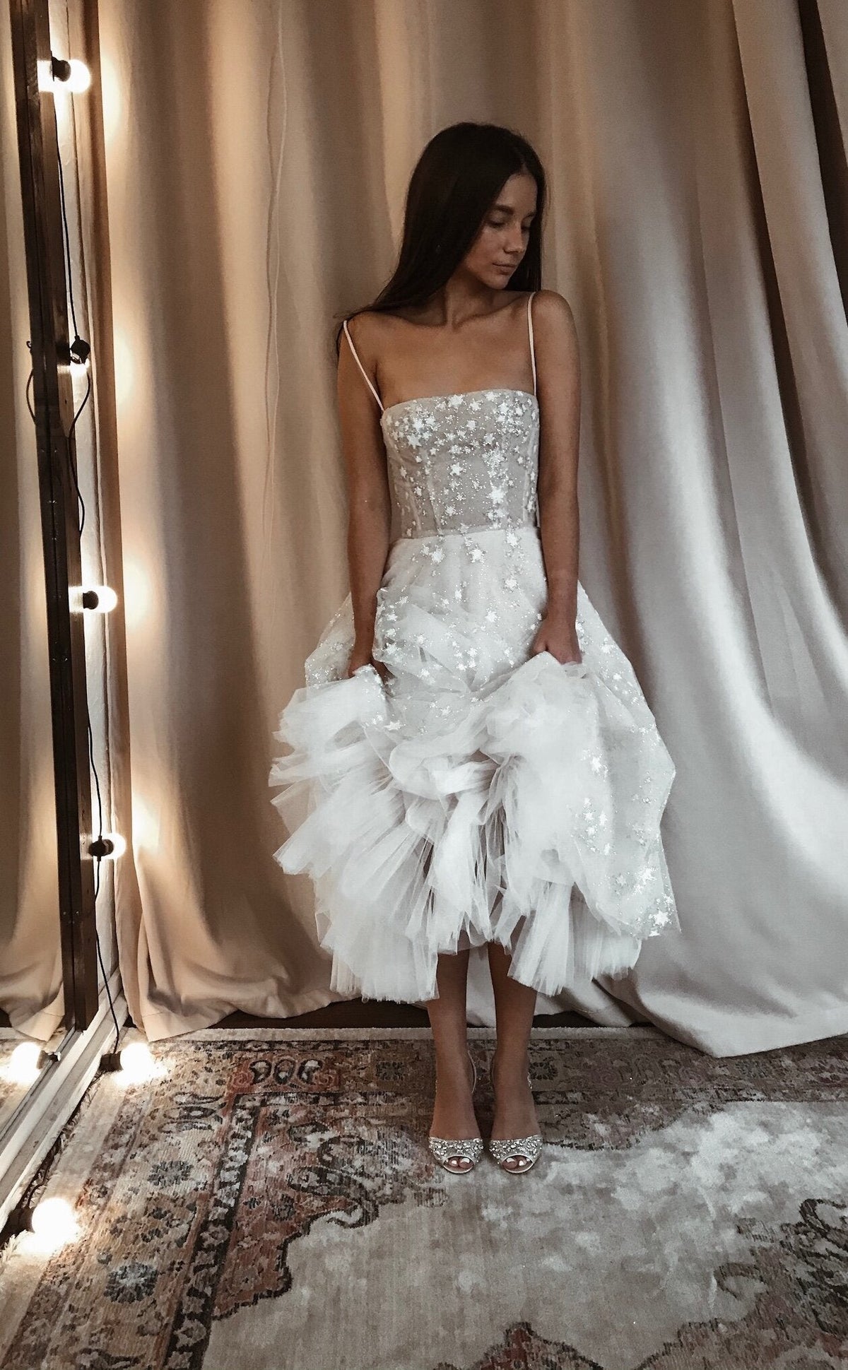 Model lifting the ruffled tulle hem of a star corset wedding dress with straps, curtain backdrop and rug, Wild Stars