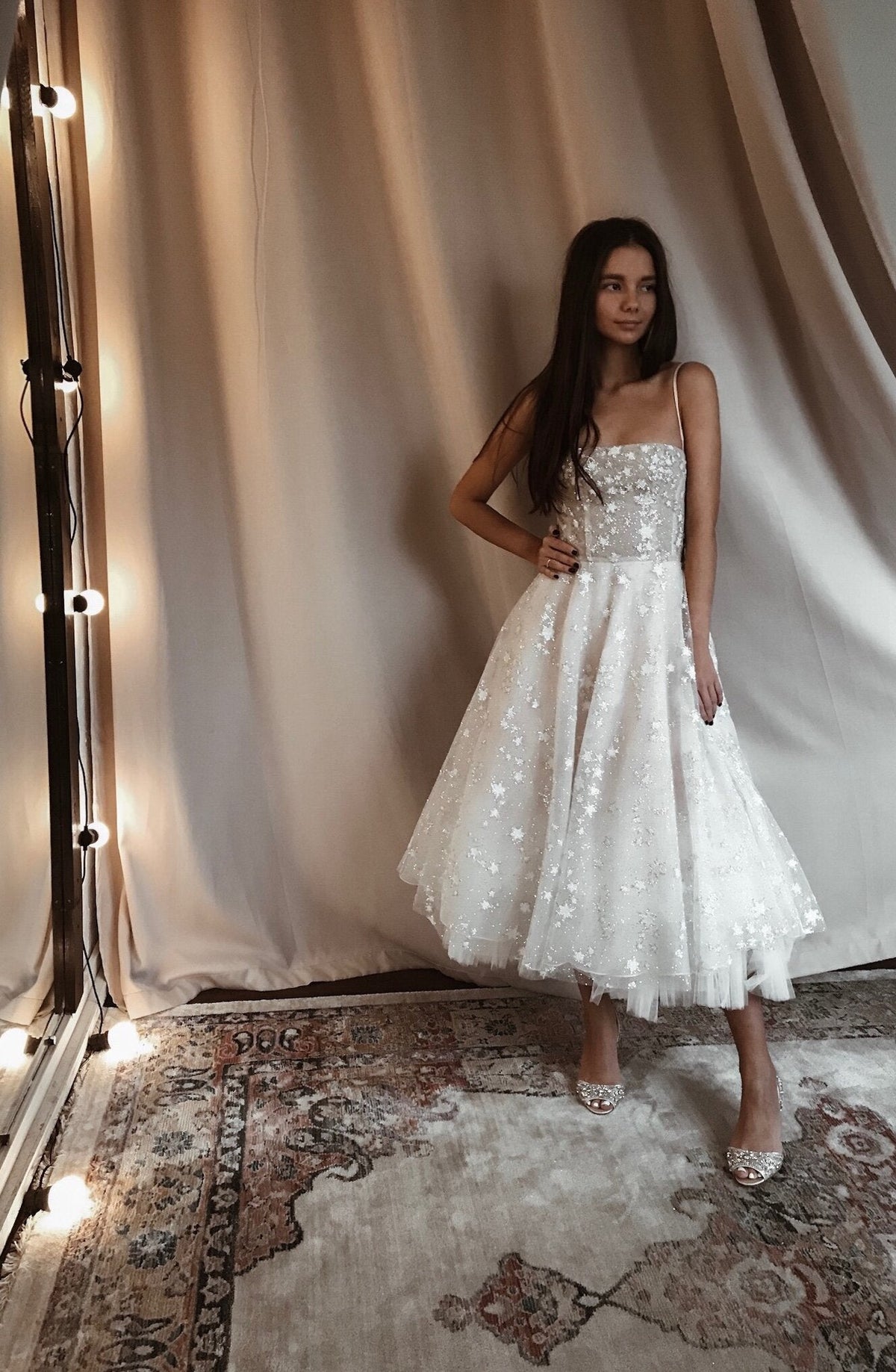Model by a curtain and bulb mirror in a tea-length star wedding dress with spaghetti straps, Persian rug, Wild Stars