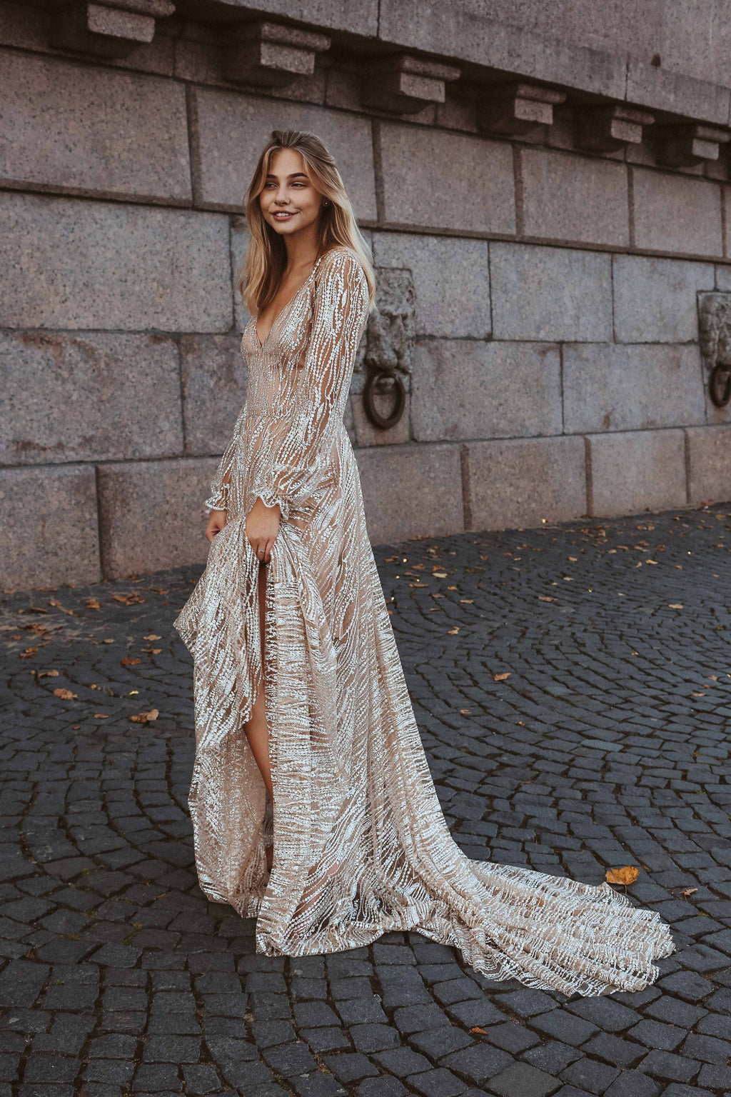 Smiling bride on a cobbled street showing the long train and front slit of a beige sparkle wedding dress, Galaxy Champagne