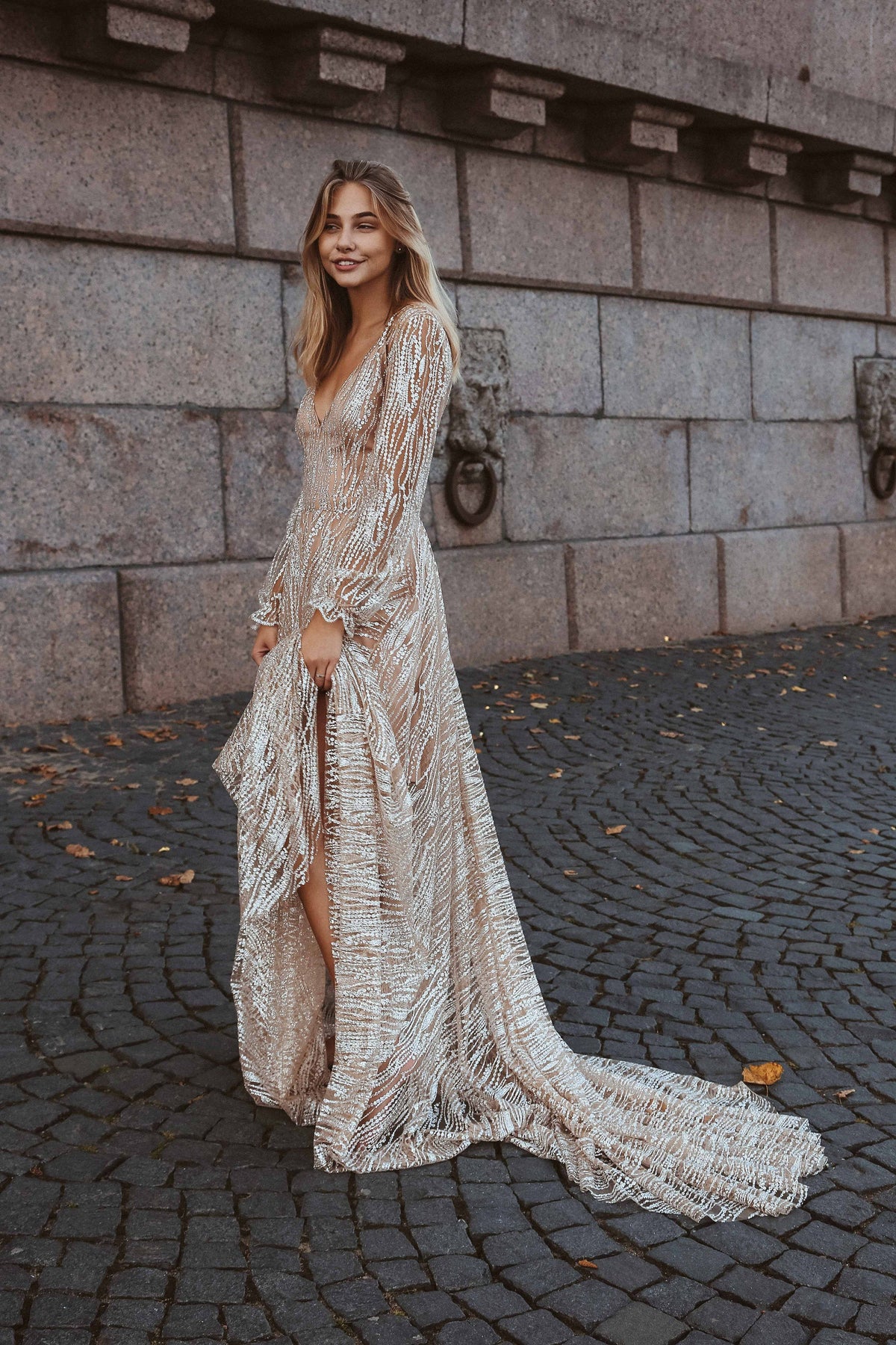 Smiling bride on a cobbled street showing the long train and front slit of a beige sparkle wedding dress, Galaxy Champagne