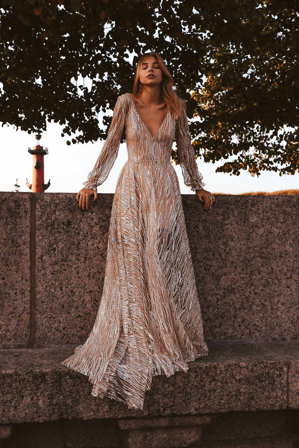 Bride on a granite embankment in a long-sleeve champagne glitter lace wedding dress with an A-line skirt, lighthouse behind, Galaxy Champagne