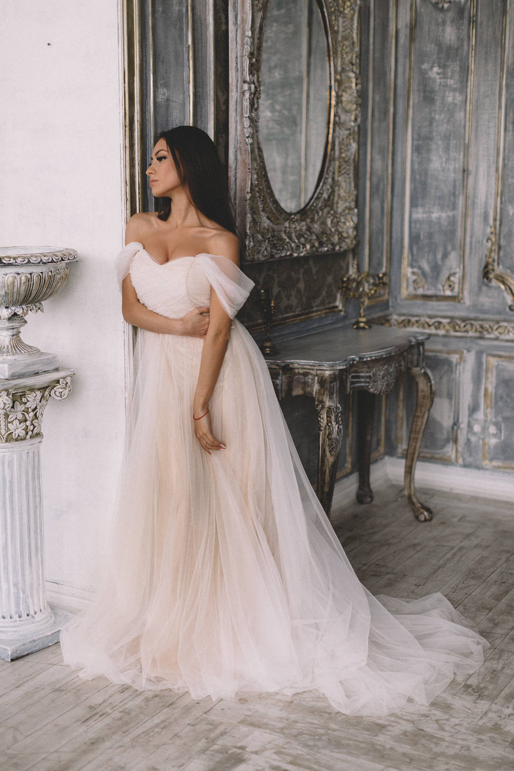 Model leaning on a pillar in a champagne tulle sample wedding dress with off-shoulder sleeves, ornate mirror behind, Whisper Sample