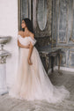 Model leaning on a pillar in a champagne tulle sample wedding dress with off-shoulder sleeves, ornate mirror behind, Whisper Sample