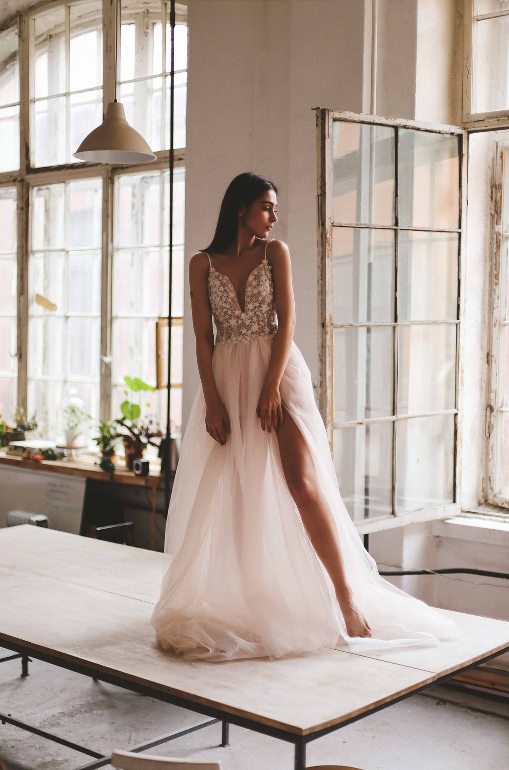 Bride seated on a long studio table by arched windows, plunge corset wedding dress with a leg slit, Make a Wish