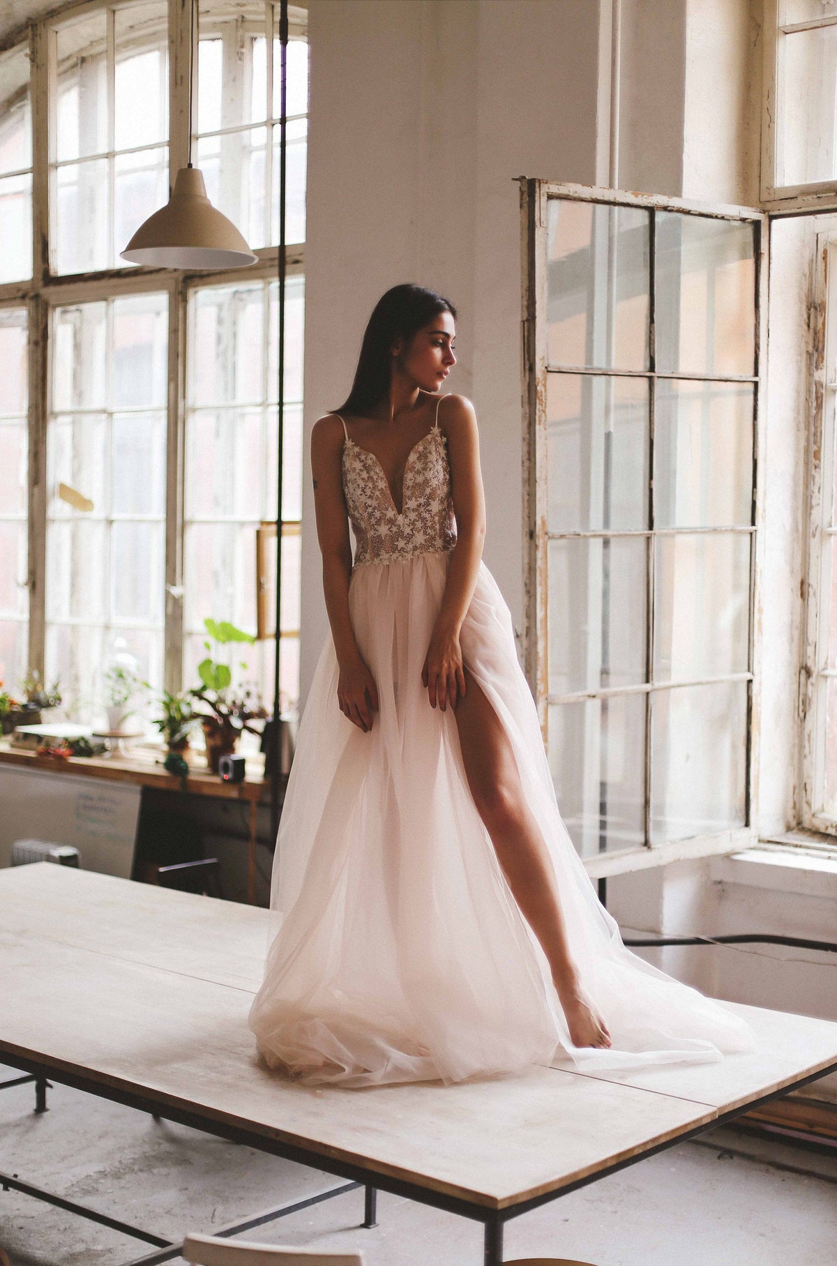 Bride seated on a long studio table by arched windows, plunge corset wedding dress with a leg slit, Make a Wish