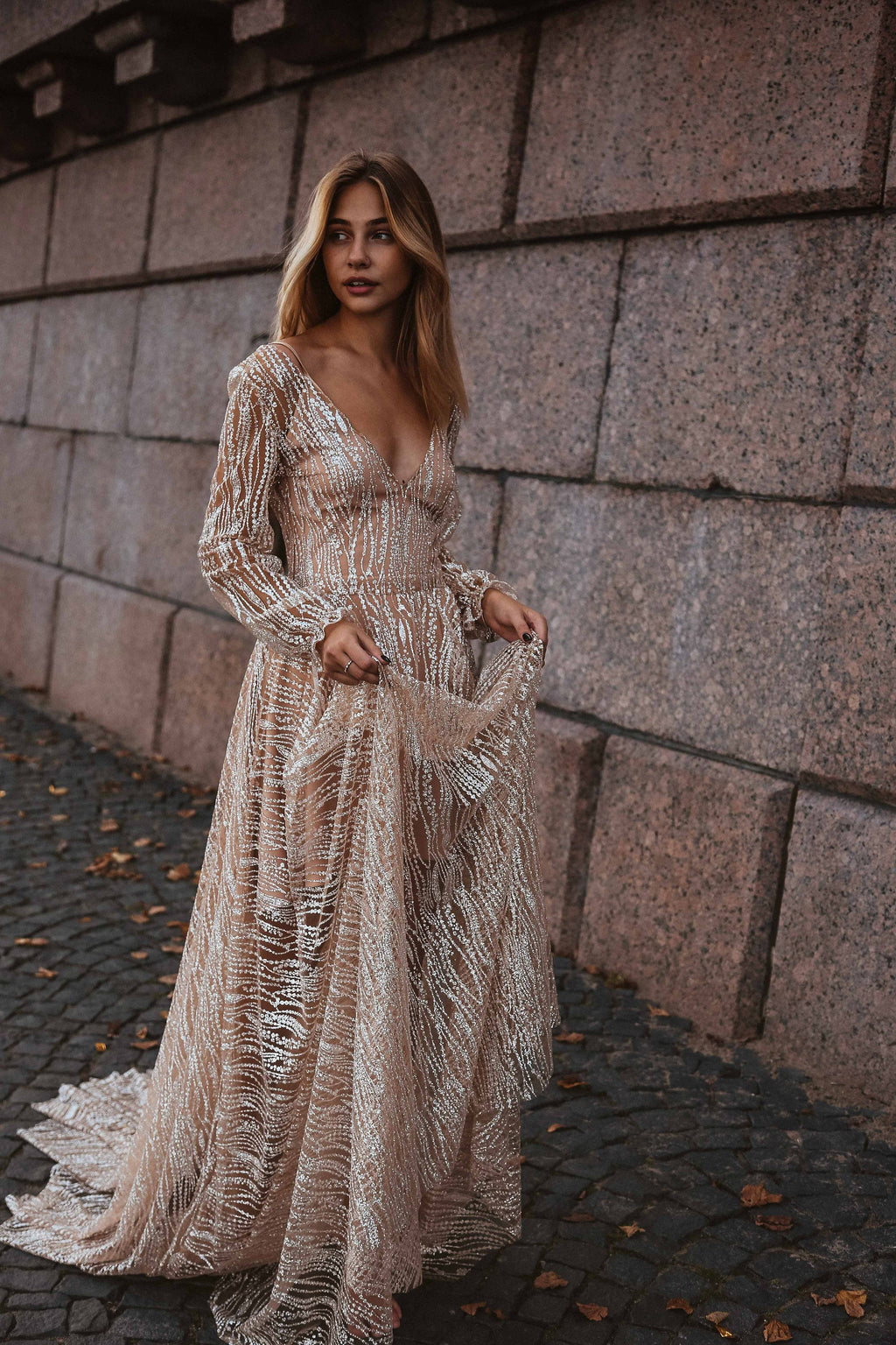 Bride lifting the skirt of a champagne lace wedding dress on cobblestones by a stone wall, train swept behind, Galaxy Champagne
