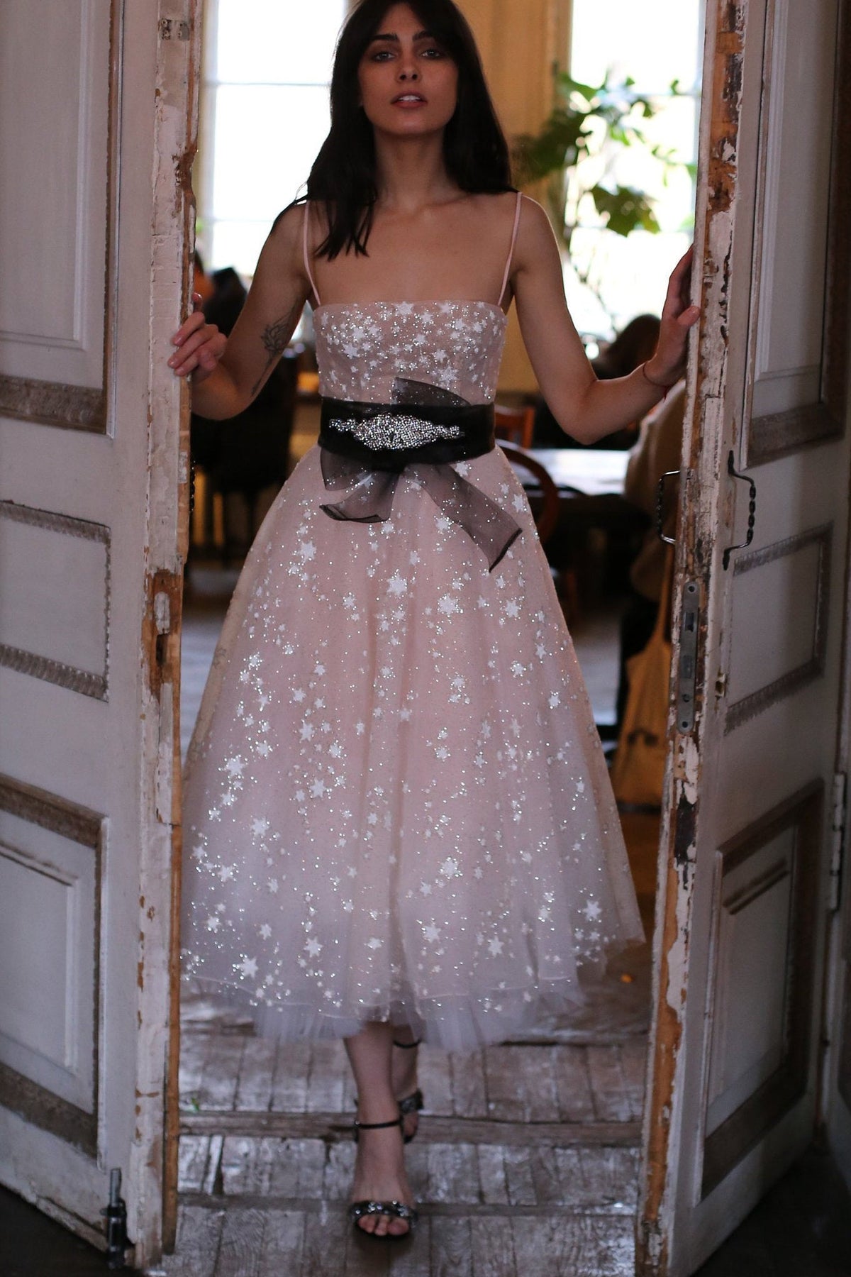 Model in a doorway in a blush star wedding dress with thin straps, black jewelled sash at the waist, Wild Stars