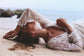 Bride lying back on the sand, arms over her head, long sheer sleeves of an ivory lace wedding dress catching the light, Galaxy Ivory