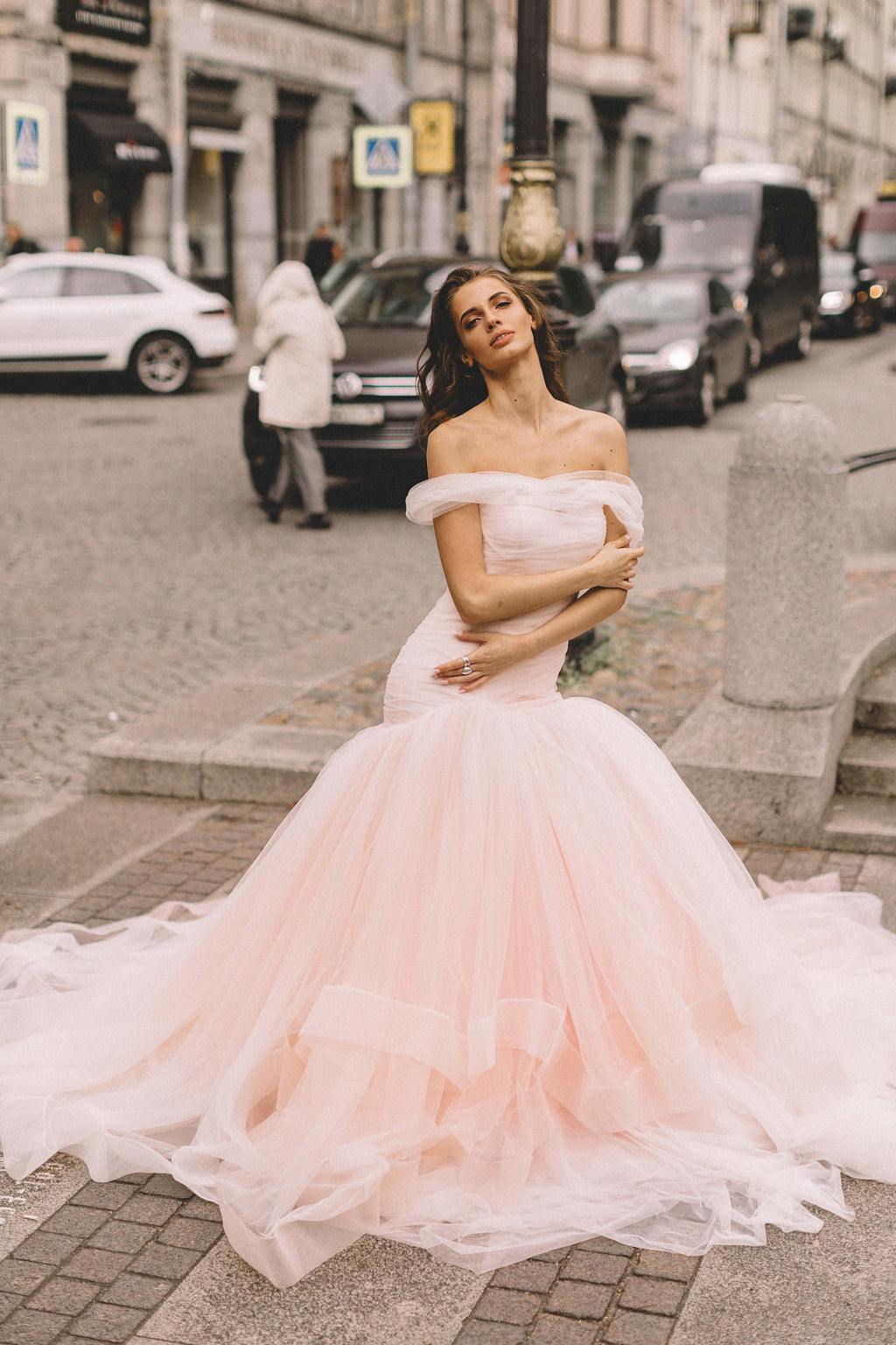Bride in a blush off-shoulder wedding dress on a city street, arms crossed, train spread over cobbles, Peony