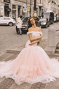 Bride in a blush off-shoulder wedding dress on a city street, arms crossed, train spread over cobbles, Peony
