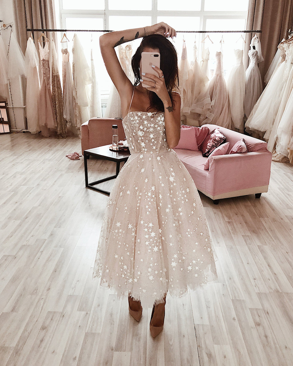 Model posing with an arm raised in a strappy midi star wedding dress, salon rails and a pink sofa behind, mirror selfie, Wild Stars