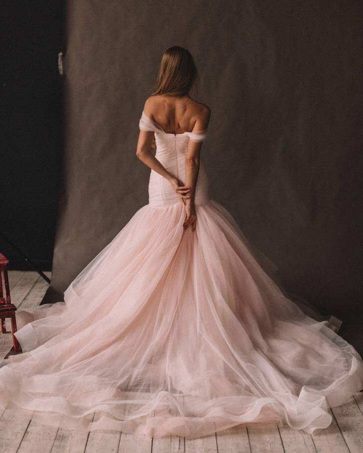 Back view of a blush mermaid wedding dress with hands clasped behind, long tulle train on wooden boards, Peony