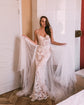 Bride against a white tiled wall holding out a tulle overskirt over a fitted lace wedding dress, Feathers Sample