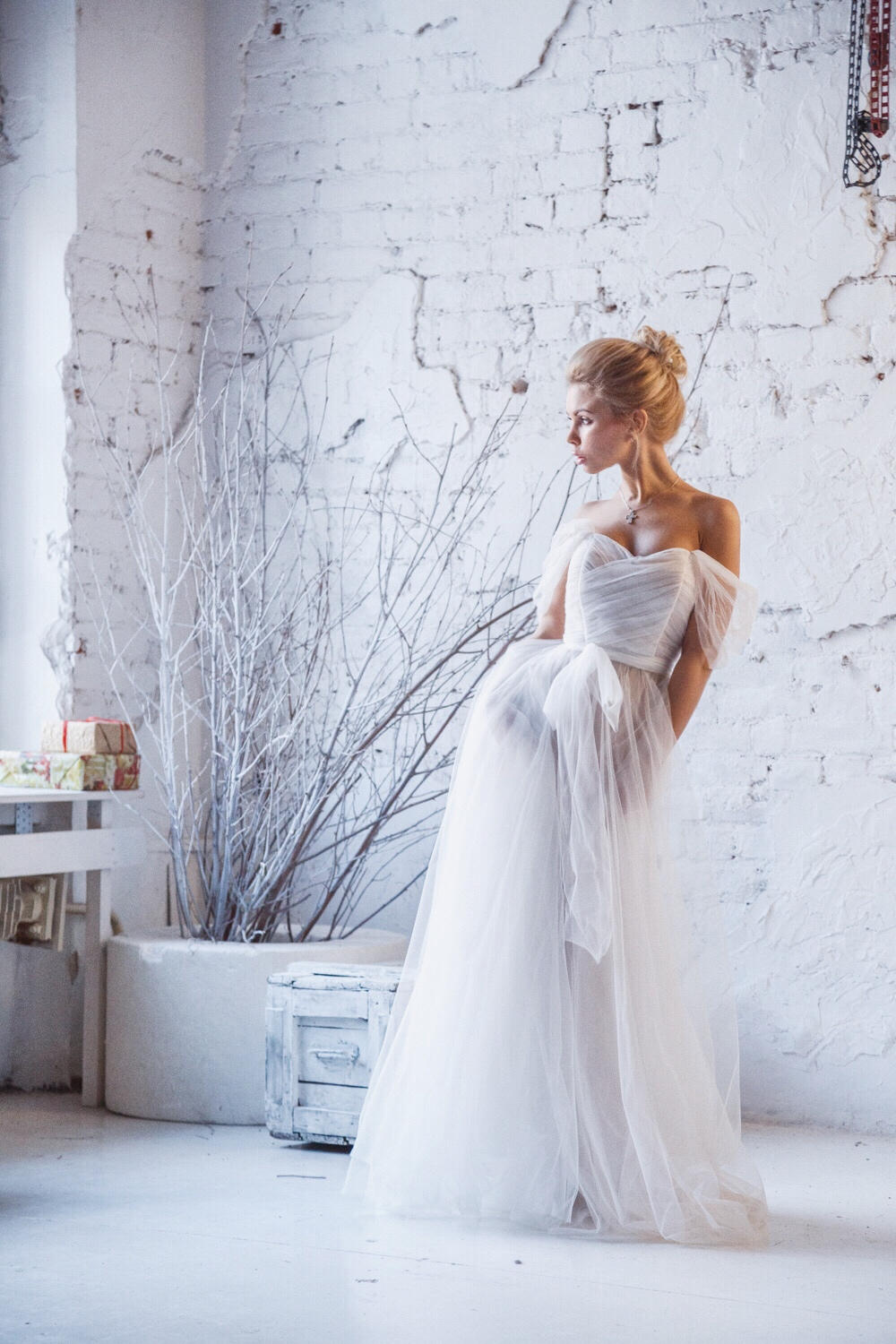 Model by a white brick wall in an off-shoulder tulle sample wedding dress with a draped pleated bodice, bare branches behind, Whisper Sample