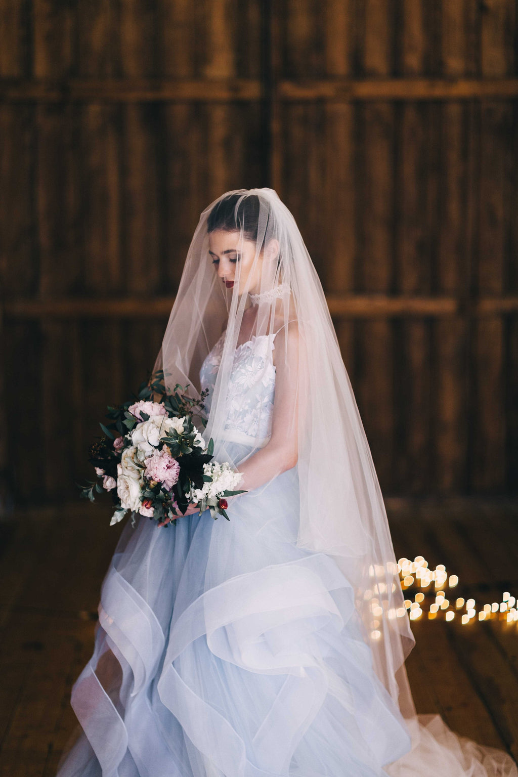 Bride looking down at her bouquet under a sheer two-tier wedding veil, front tier over the face, fairy lights behind, Cascade Veil
