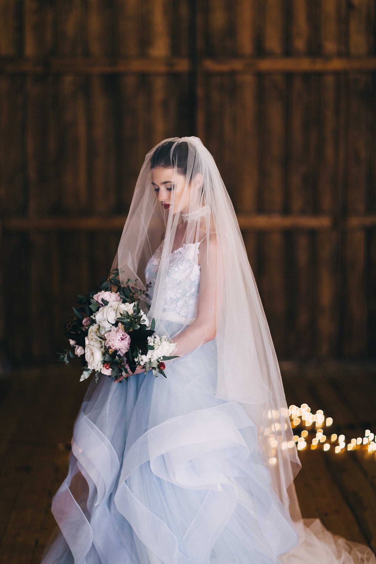 Bride looking down at her bouquet under a sheer two-tier wedding veil, front tier over the face, fairy lights behind, Cascade Veil