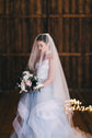 Bride looking down at her bouquet under a sheer two-tier wedding veil, front tier over the face, fairy lights behind, Cascade Veil